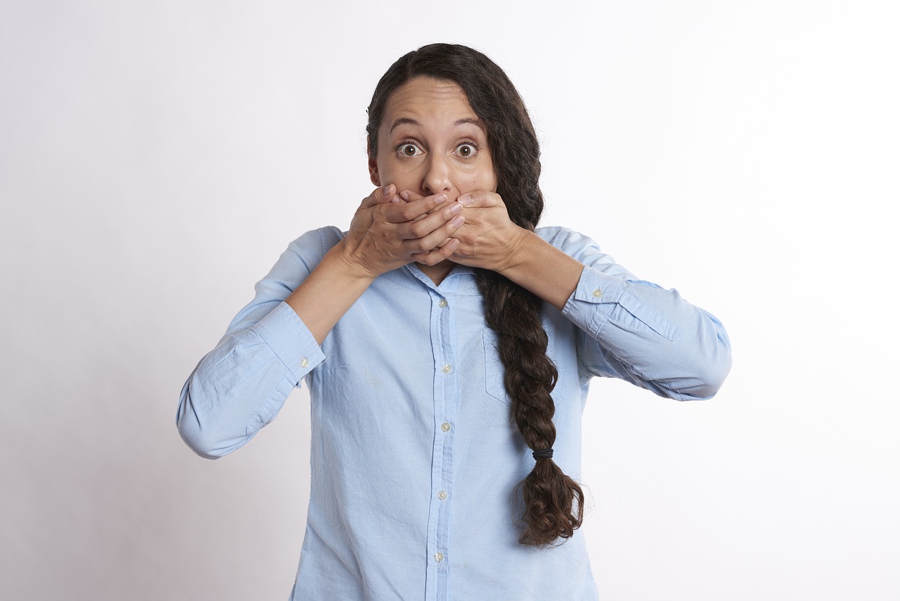 Surprised woman with wide eyes covering her mouth with both hands, wearing a light blue button-up shirt, standing against a plain white background.